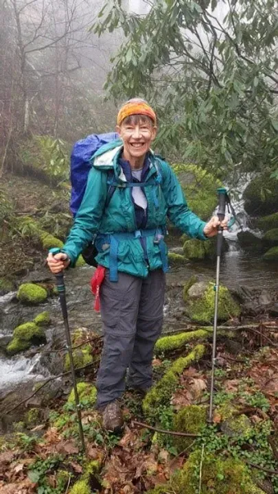 A woman with a backpack and hiking poles is standing next to a stream.