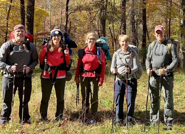 A group of people standing next to each other in the woods holding hiking poles.