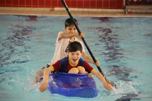 Two young boys are rowing a kayak in a swimming pool.
