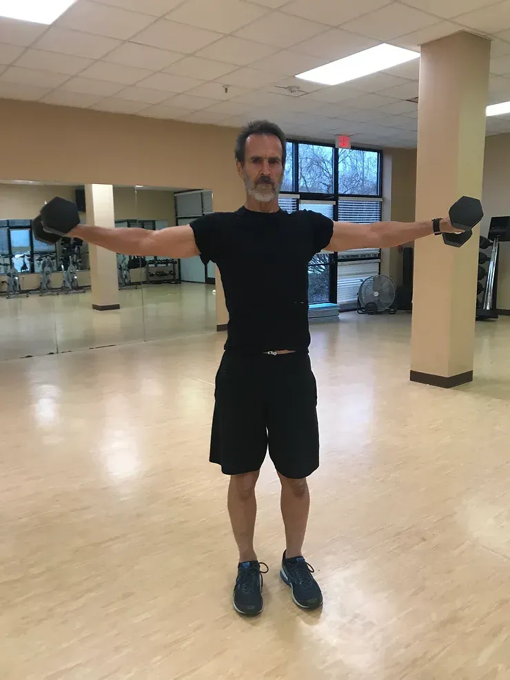 A man is holding a dumbbell in a gym with his arms outstretched.
