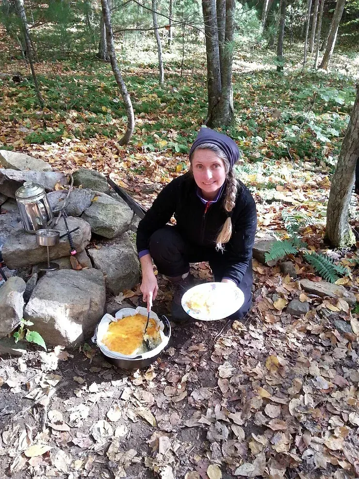 A woman is kneeling down in the woods with a bowl of food.