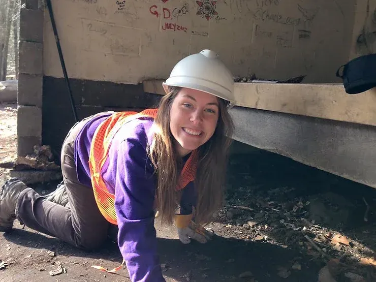 A woman wearing a hard hat and an orange vest is kneeling on the ground.
