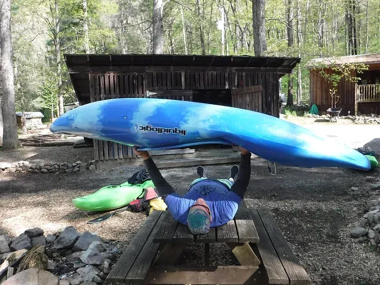 A man is laying on a picnic table holding a blue kayak over his head.