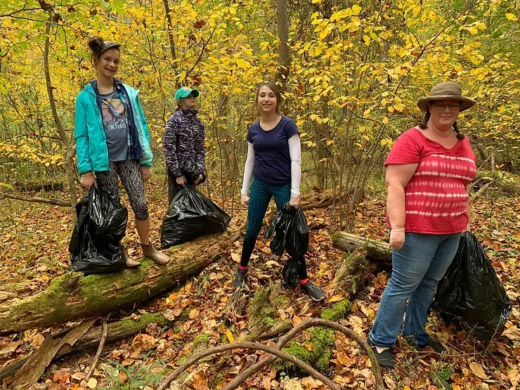 A group of people are standing on a log in the woods holding trash bags.