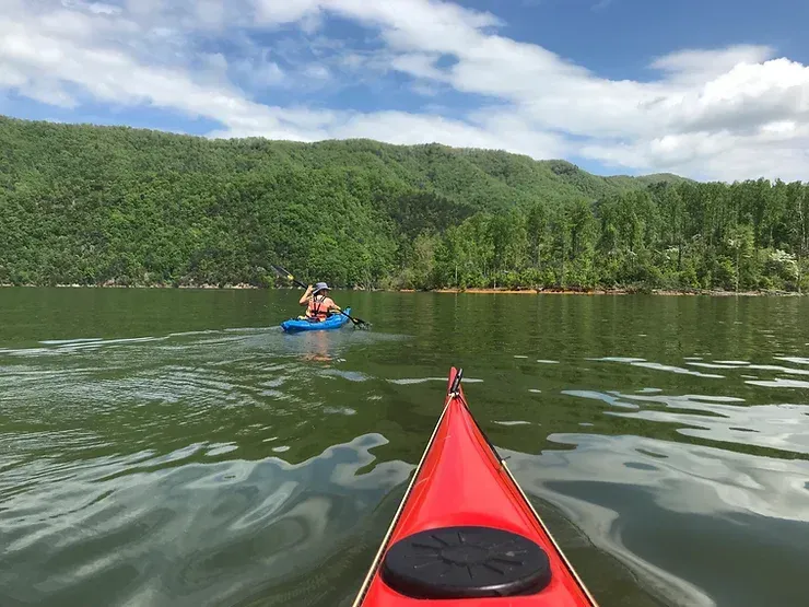 A person is paddling a kayak on a lake with mountains in the background.