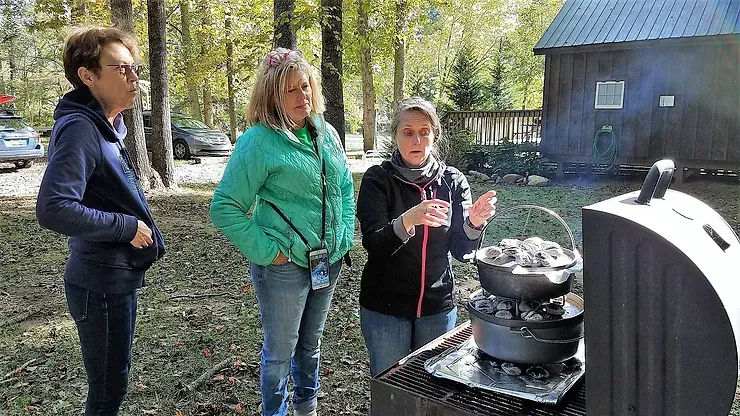 Three women are standing around a dutch oven on a grill.