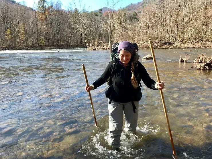 A woman with a backpack is crossing a river with sticks.