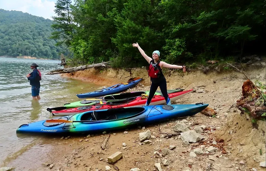 A woman is standing on top of a kayak on the shore of a lake.