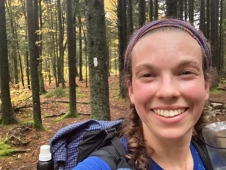 A woman with a backpack is smiling in the woods