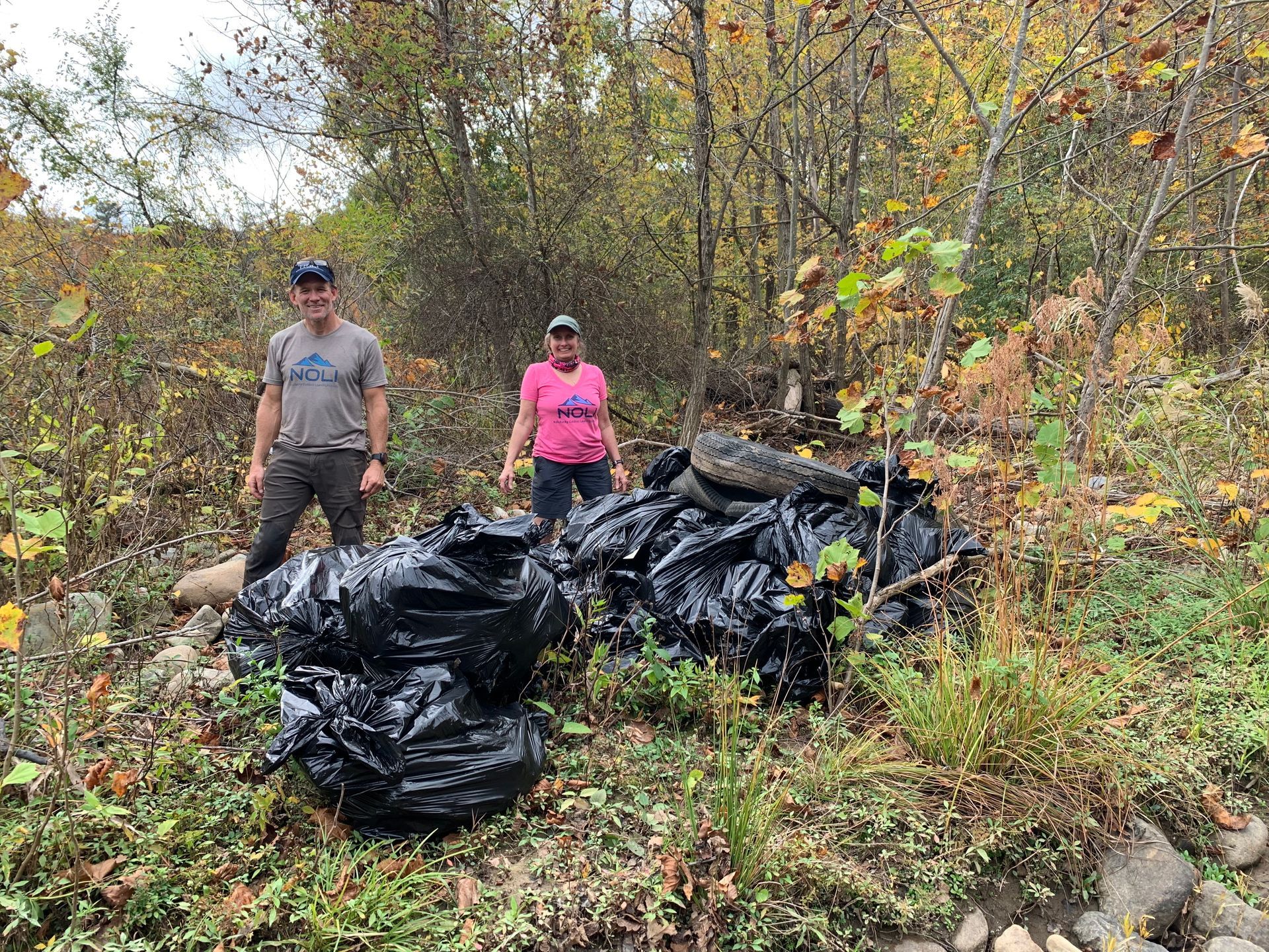 A man and a woman are standing next to a pile of trash bags in the woods.