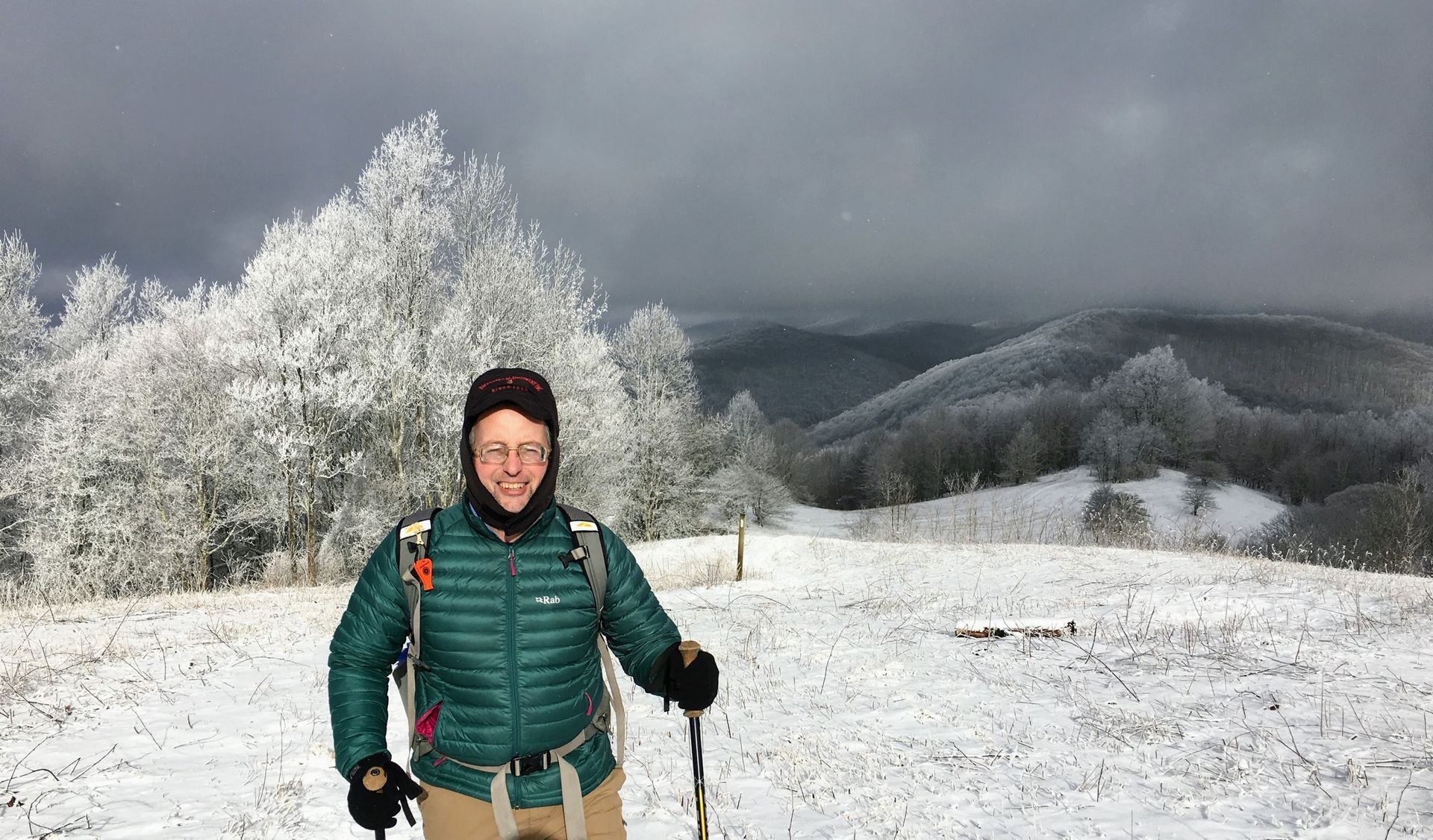 A man in a green jacket is standing in the snow with hiking poles.