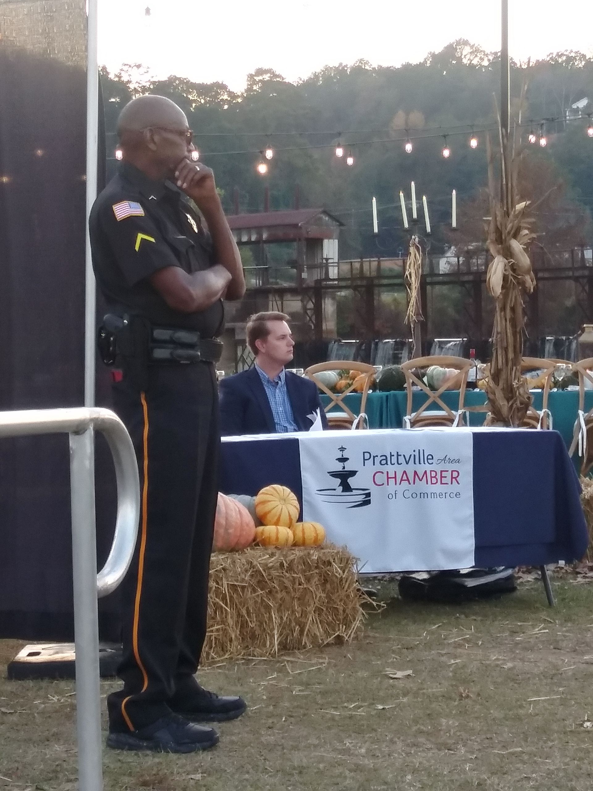 Man in black uniform speaks at outdoor cancer fundraiser beside hay bales and a table with a banner