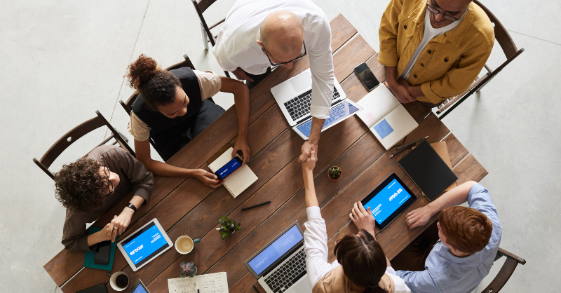 Top-down view of a team meeting around a wooden table with laptops, notebooks, and tablets
