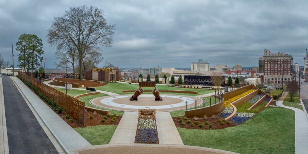 Landscape park with a circular fountain, paths, flower beds, and buildings under a cloudy sky