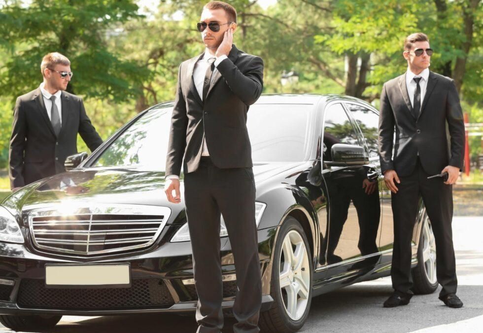 Three suited bodyguards standing by a black luxury sedan on a sunny street