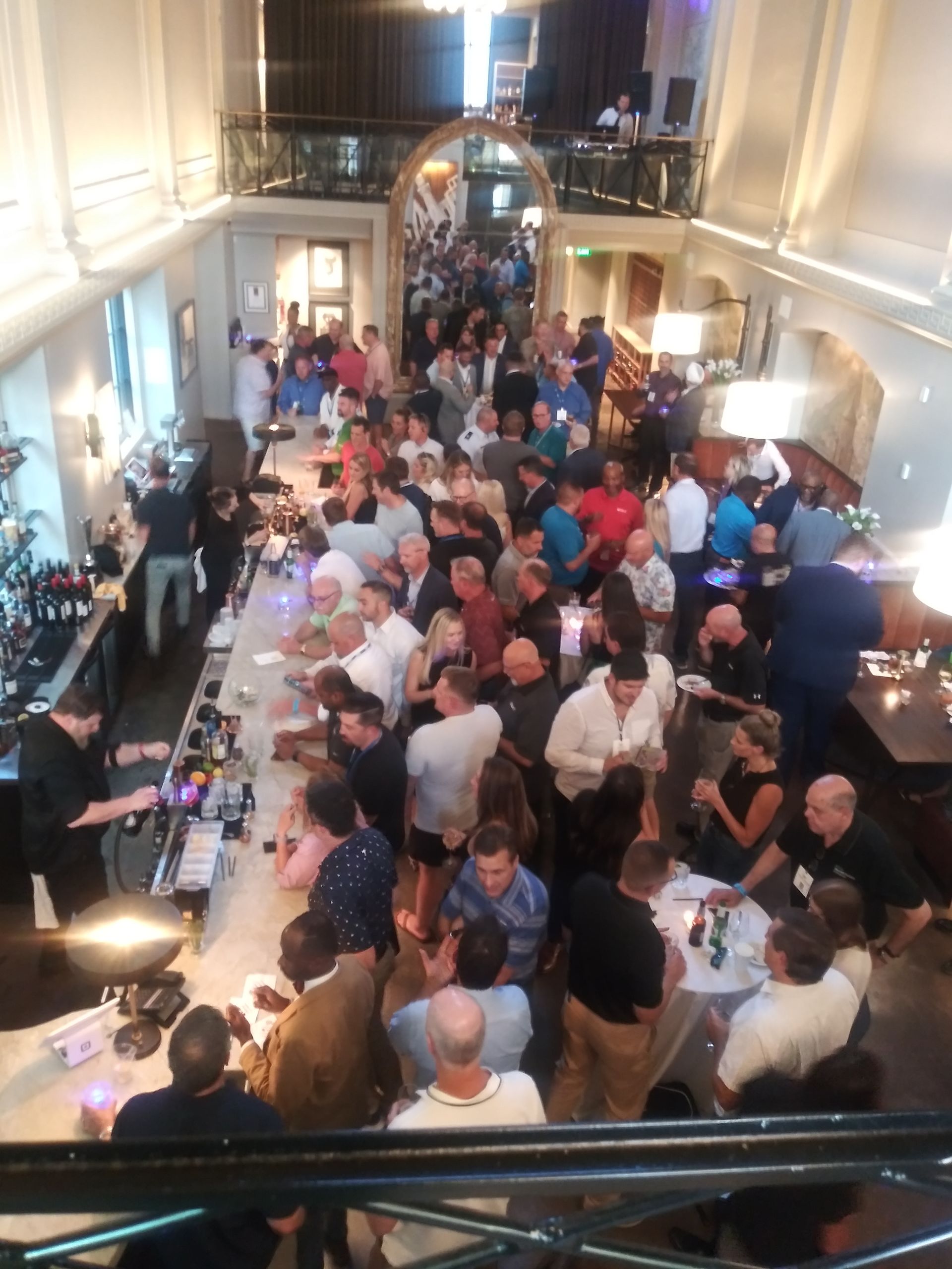 Crowded reception in a bright hall, with guests mingling around long white-clothed tables and a bar.