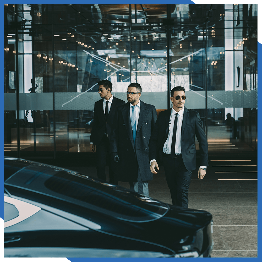 Three suited men walk past a sleek black car outside a glass-fronted building.