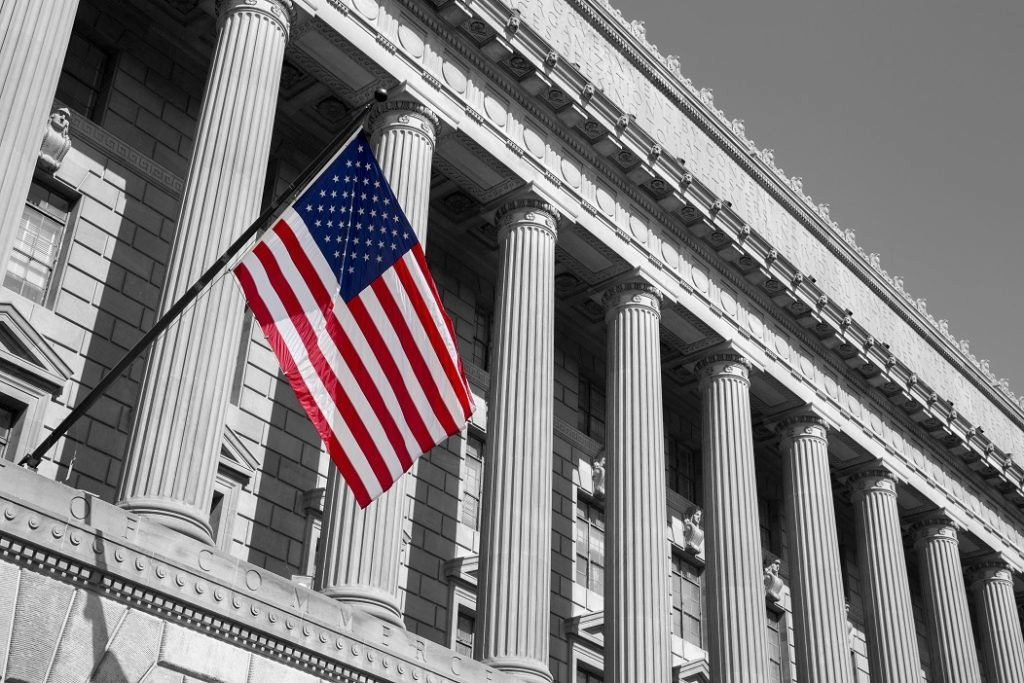 American flag hanging from a neoclassical building with columns, shown in grayscale with red, white, and blue flag colors