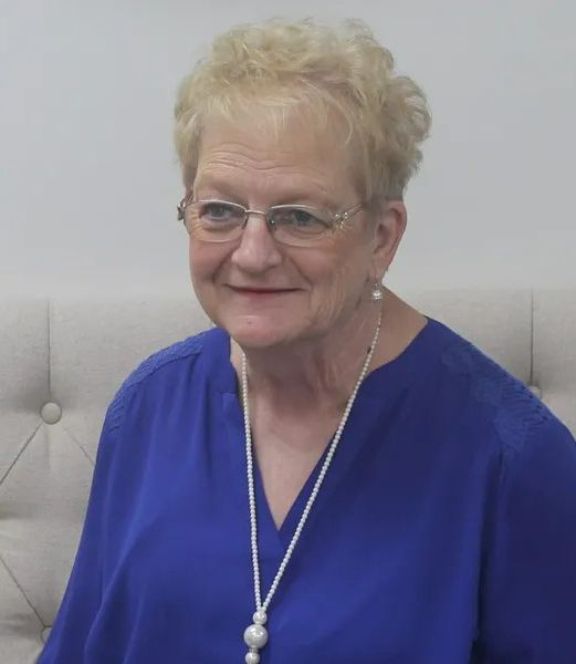 Woman in blue blouse with glasses and pearl necklace, smiling against a light tufted wall.
