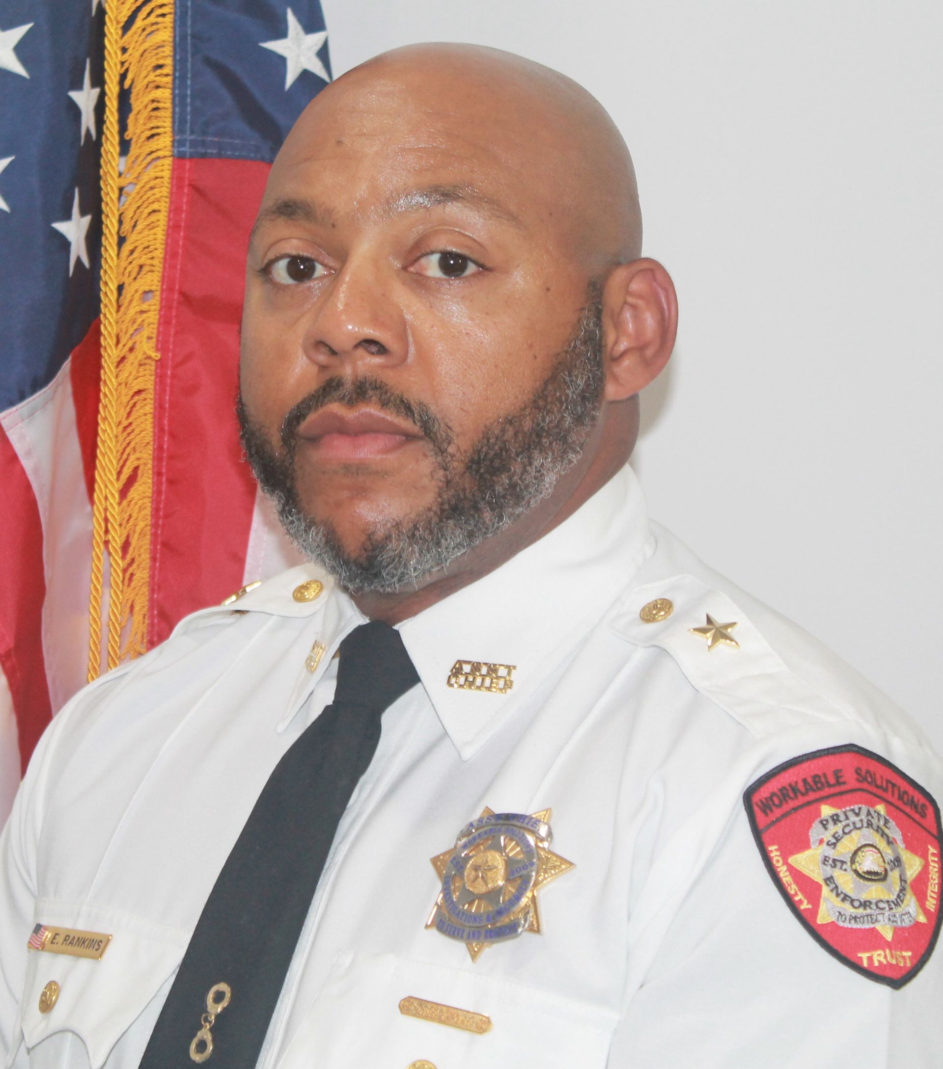 Uniformed officer standing beside a U.S. flag, wearing a white dress shirt and looking at the camera