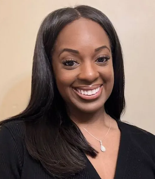 Smiling woman with long black hair in a black top, wearing a silver pendant necklace, against a beige background