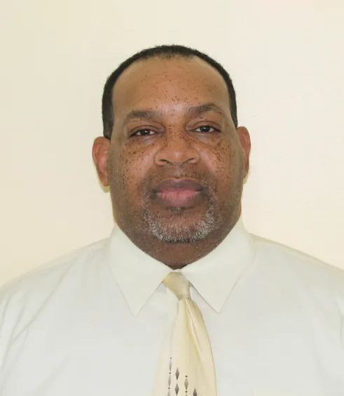 Studio portrait of a man in a white shirt and pale tie, facing forward against a plain beige background