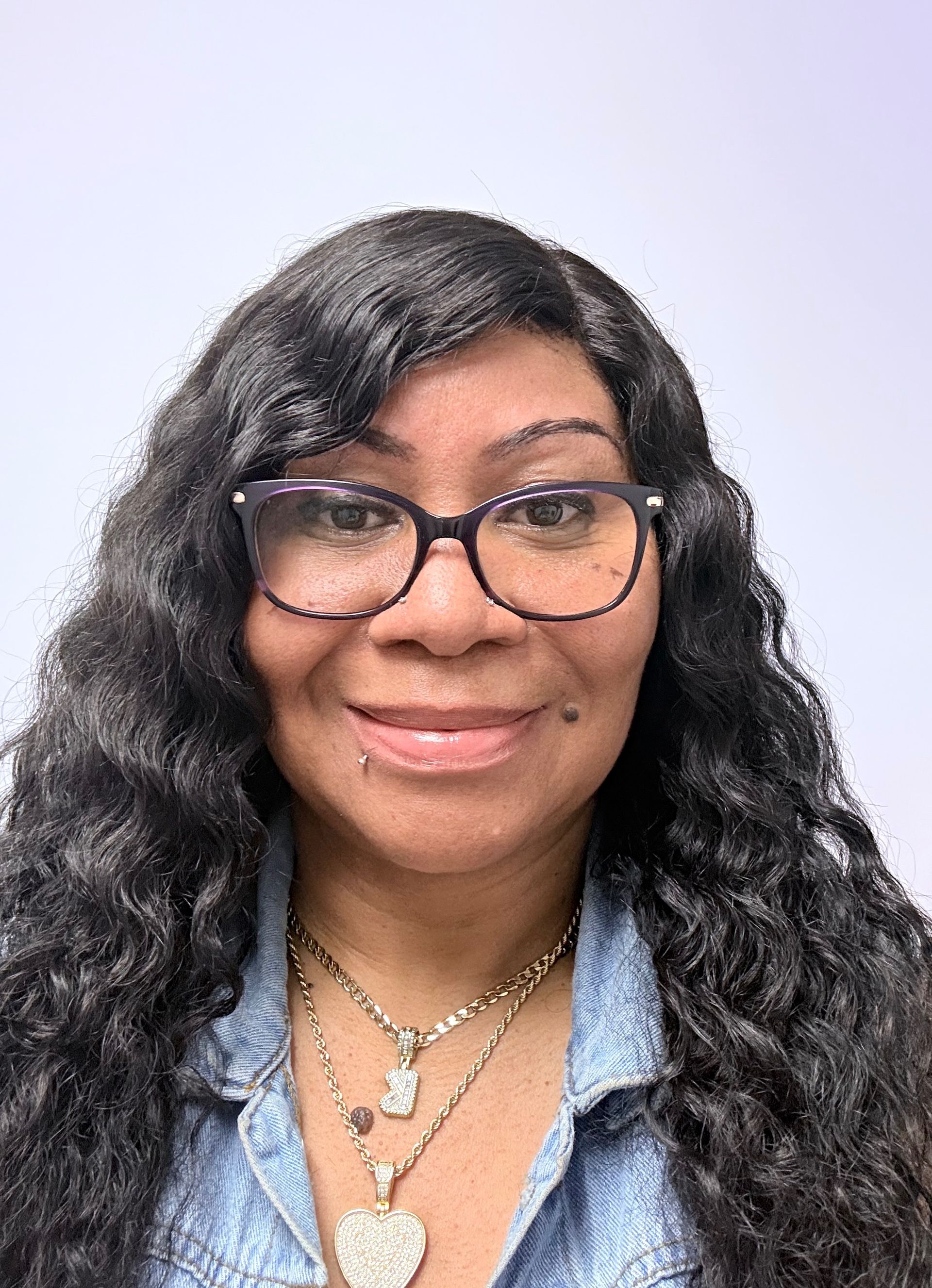 Smiling person with glasses and long wavy hair wearing a denim shirt and layered necklaces against a light background