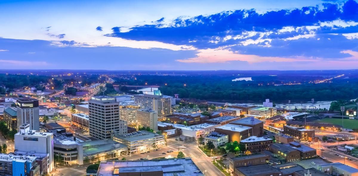 Aerial view of a city skyline at dusk with lit buildings, roads, and a blue sky over the horizon