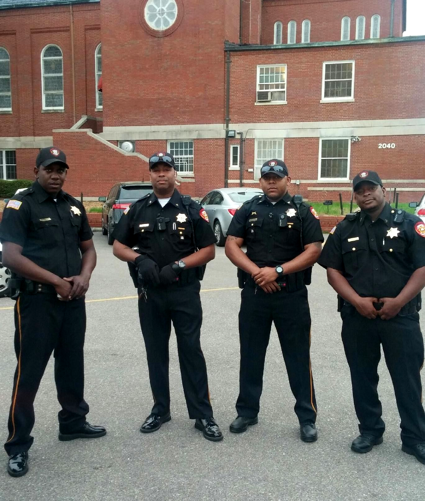 Four police officers standing in front of a brick building and parked cars
