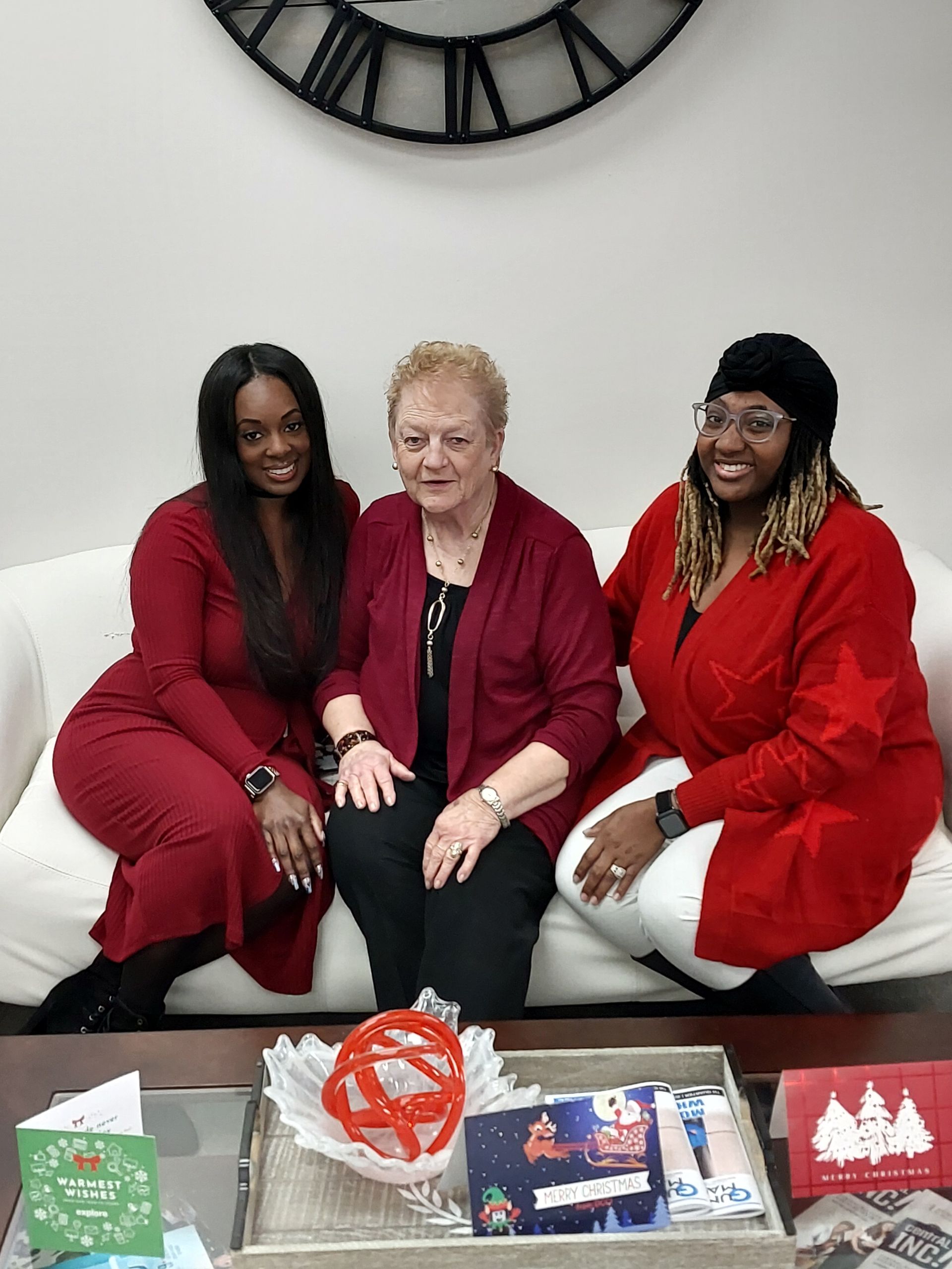 Three people seated on a white couch in red outfits, smiling with gifts on a table in front of them.