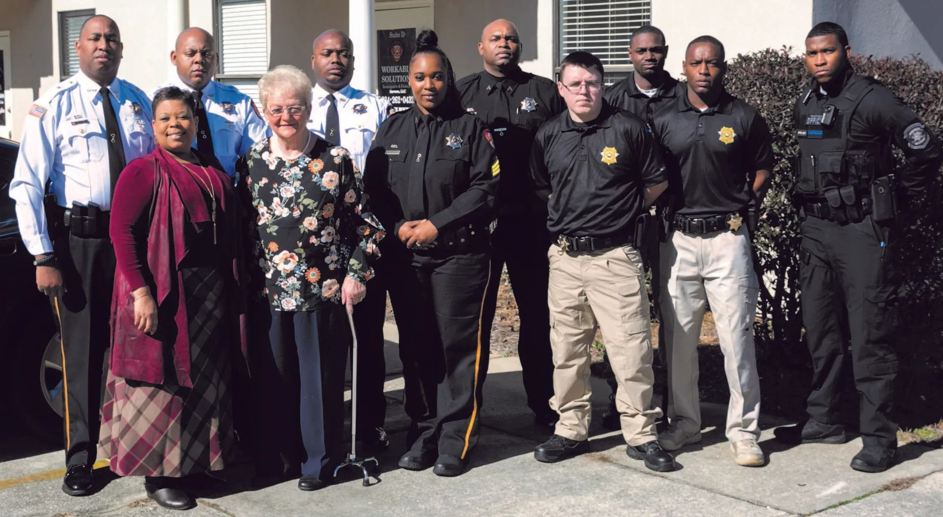 Group of people posing outdoors in front of a building, several in black shirts and beige pants.