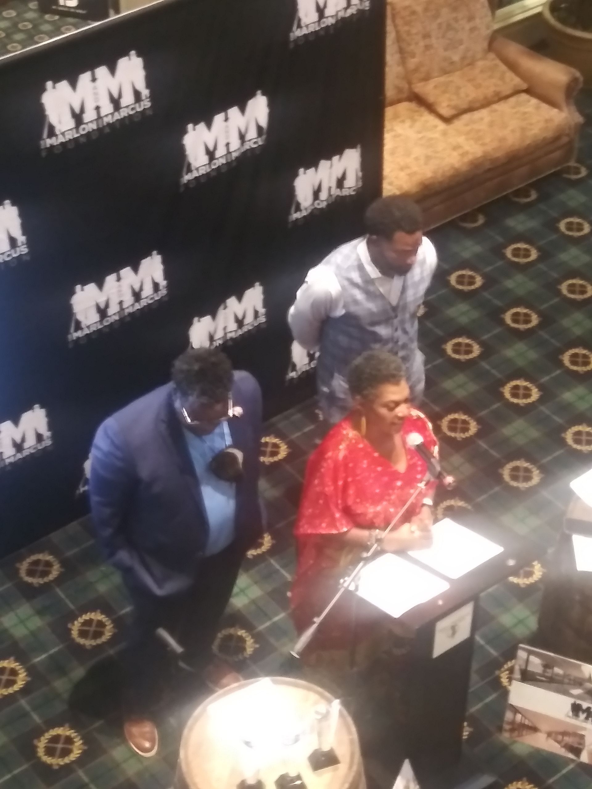 Three people stand at a podium beside a backdrop with repeated “MM” logos, viewed from above.