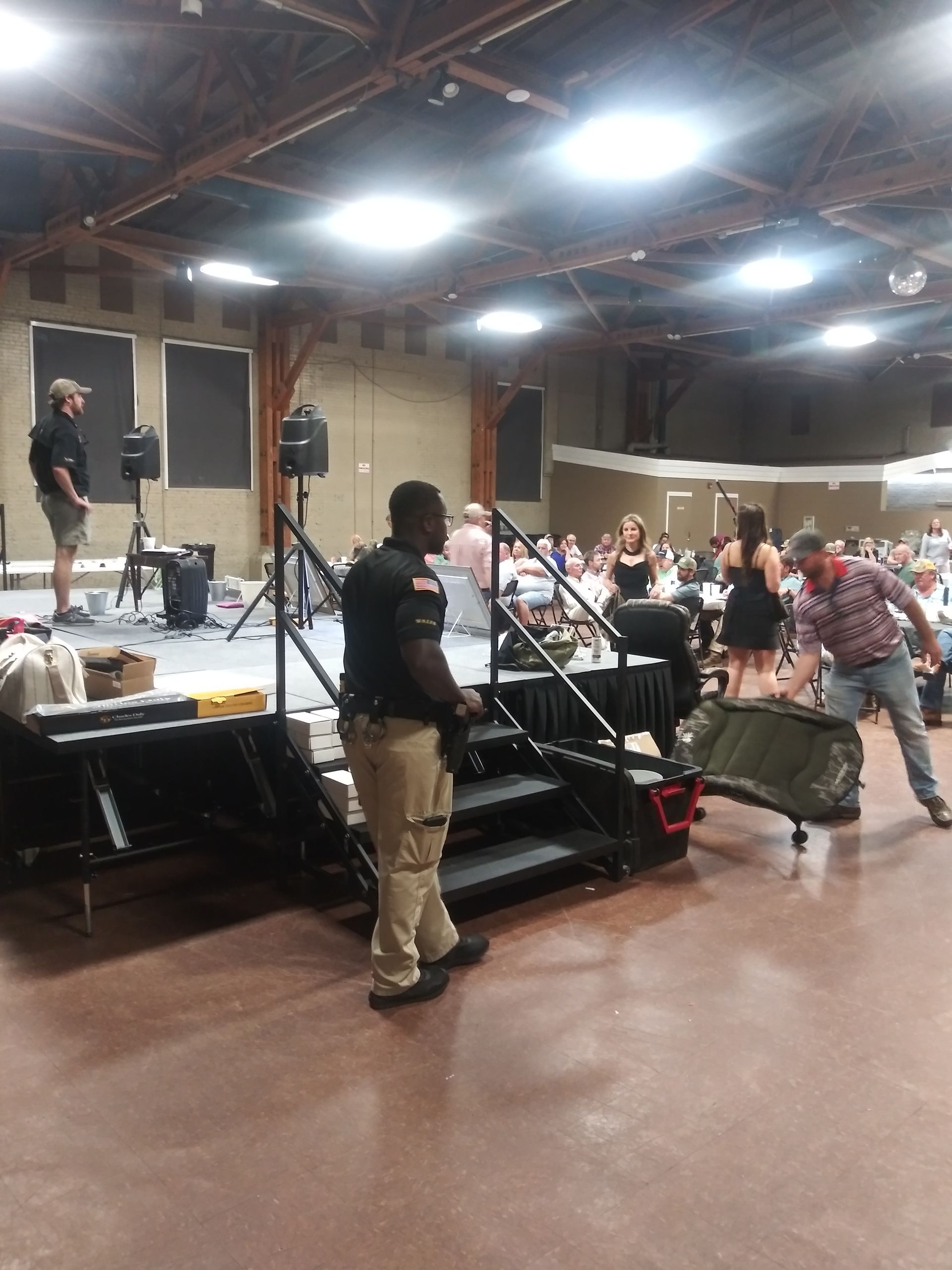 Musicians setting up on a small stage in a hall, with microphones, drums, and a person carrying equipment.