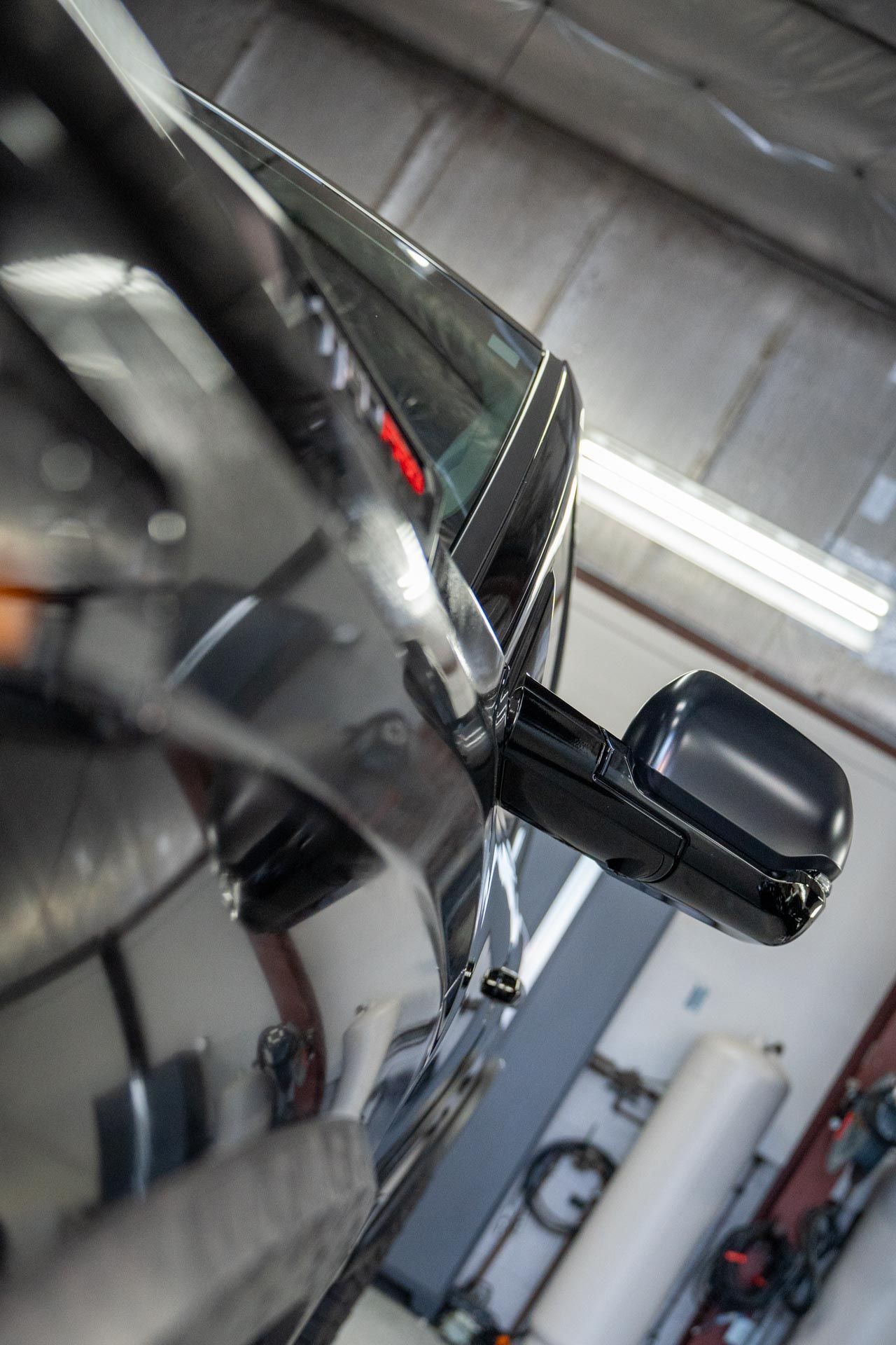 Side view of a shiny black car parked in a garage, with a side mirror and workshop equipment nearby