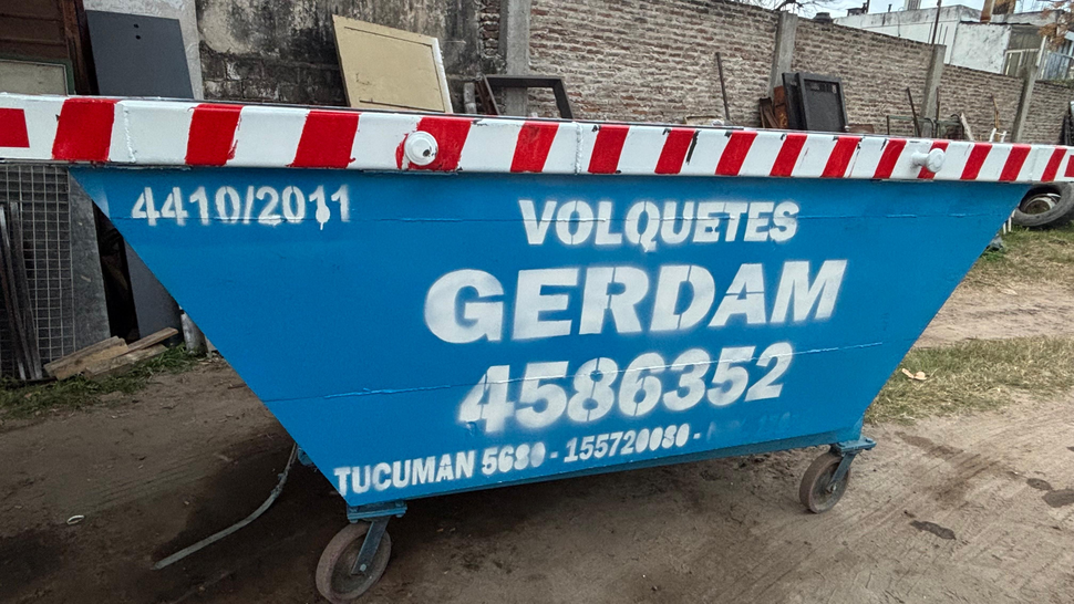 Gran contenedor de basura gris en una obra de construcción con trabajadores y maquinaria al fondo.