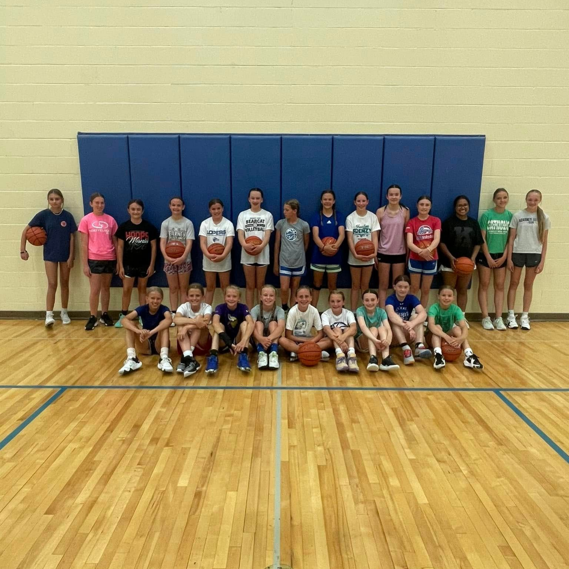 Group of children posing in a gym, some standing and some kneeling, with basketballs and blue wall pads behind them.