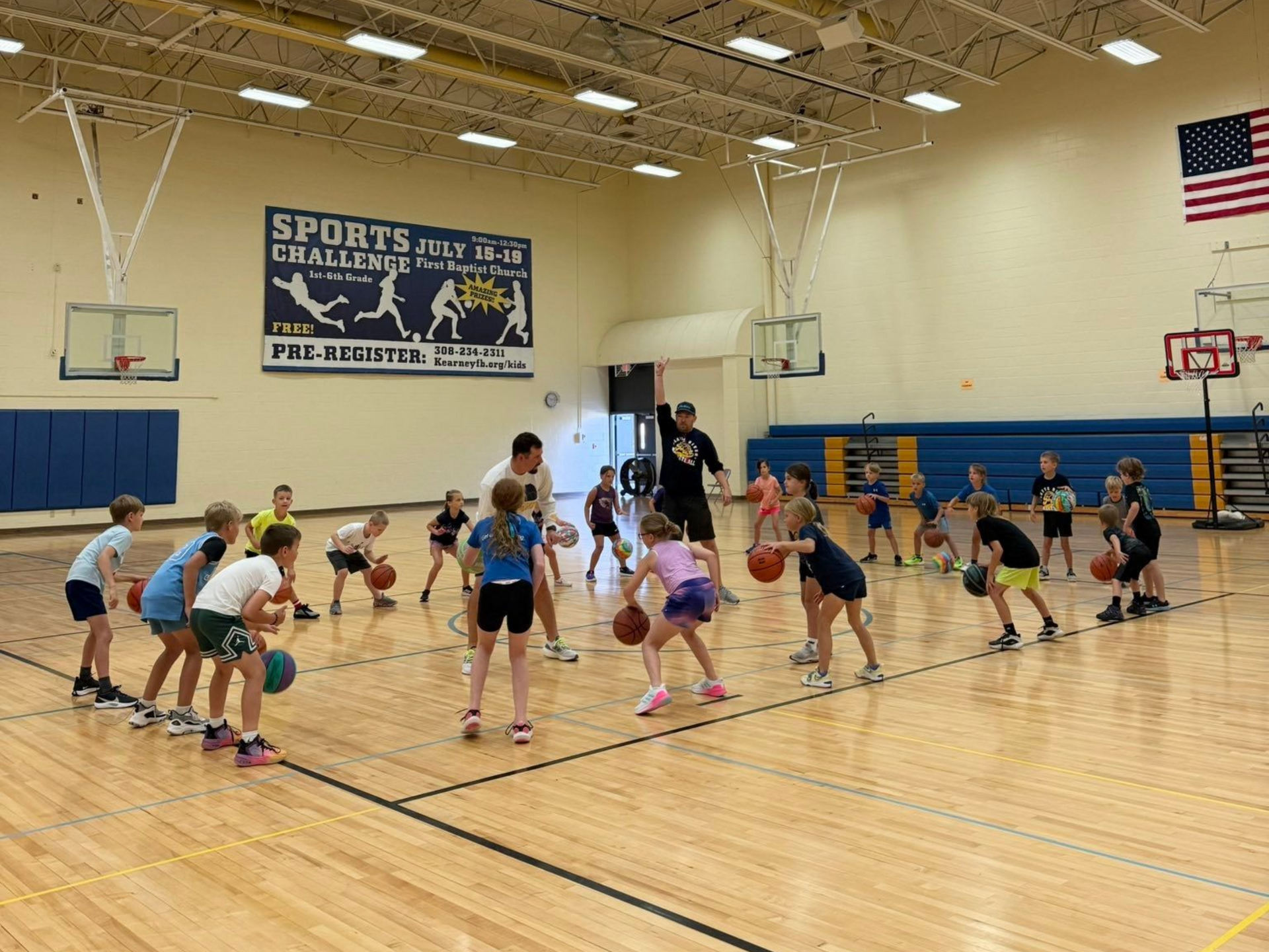 Children stretching in a gym during a basketball practice, with hoops, bleachers, and an American flag visible.