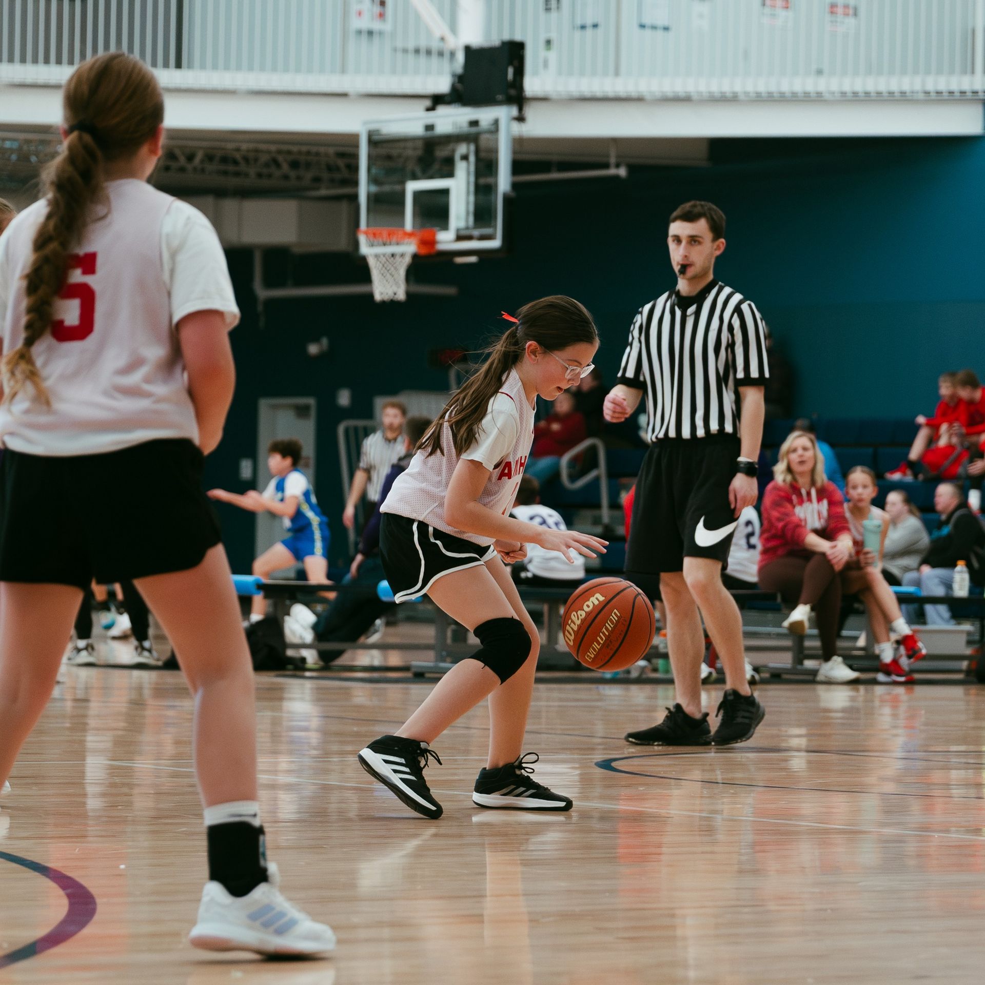Youth basketball game in a gym, with a player dribbling past a referee and spectators in the background