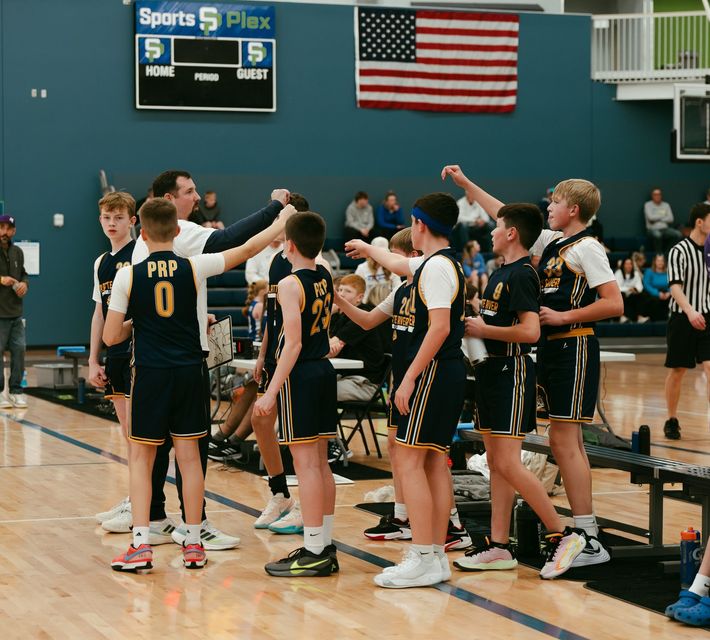 Youth basketball team huddled on a gym court during a game, with an American flag and scoreboard in the background