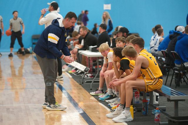 Youth basketball coach talks to players seated on a bench during a game in a gymnasium.