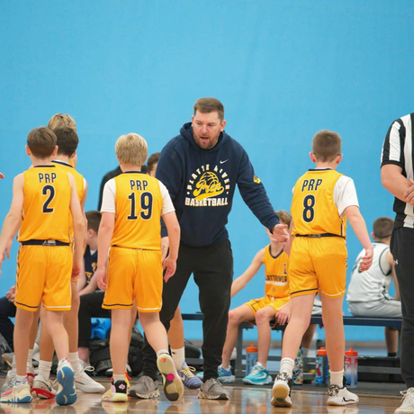 Youth basketball coach kneels courtside, talking to players in yellow jerseys during a timeout
