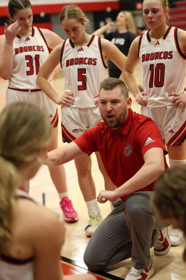 Basketball coach kneels and instructs players during a timeout on a gym floor.