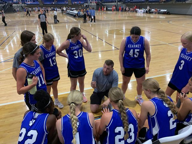 Women’s basketball team huddles around coach on a gym court, wearing blue jerseys and listening closely