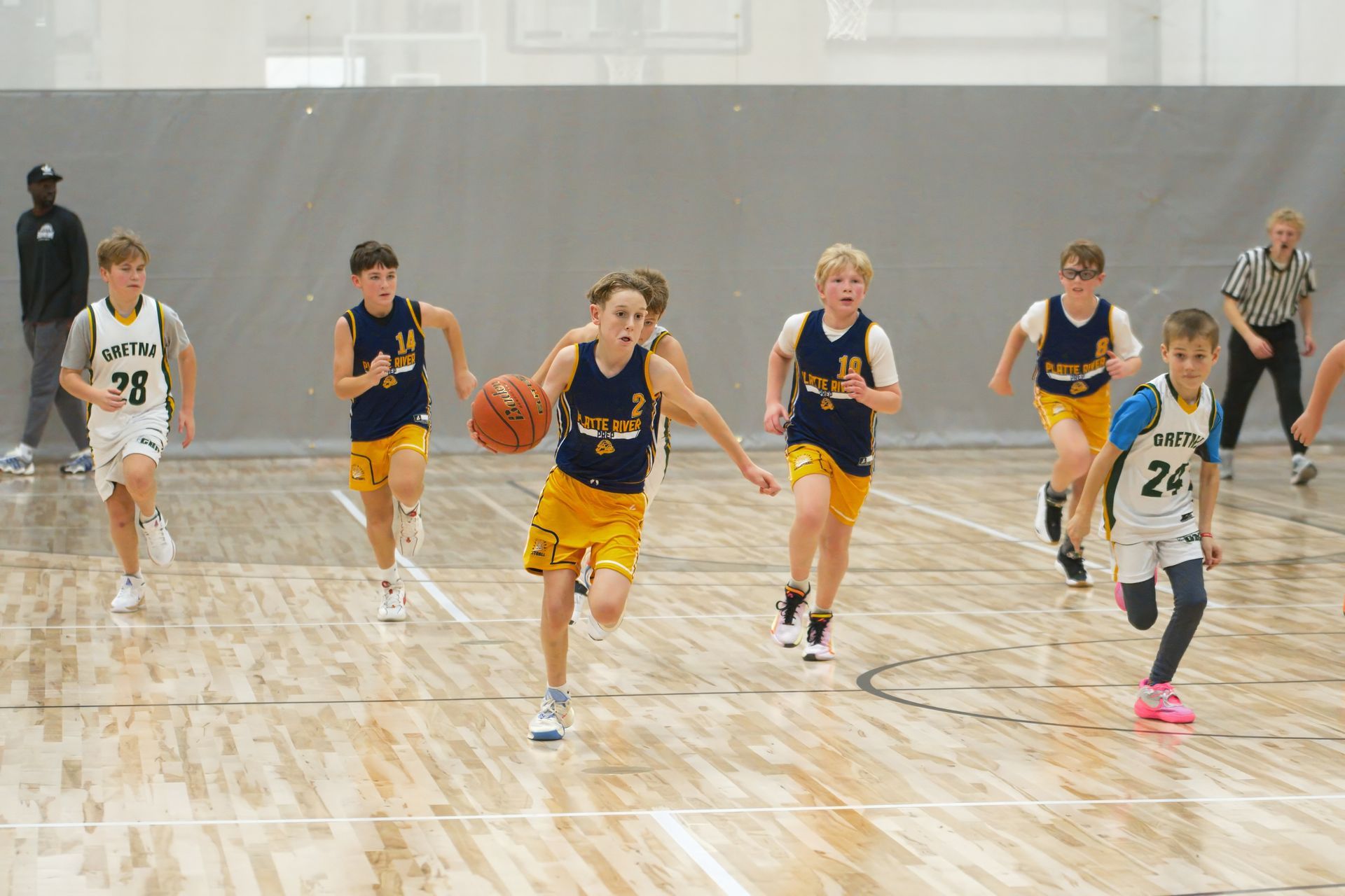 Basketball players sprint down a gym court during a game, with one dribbling toward the camera.
