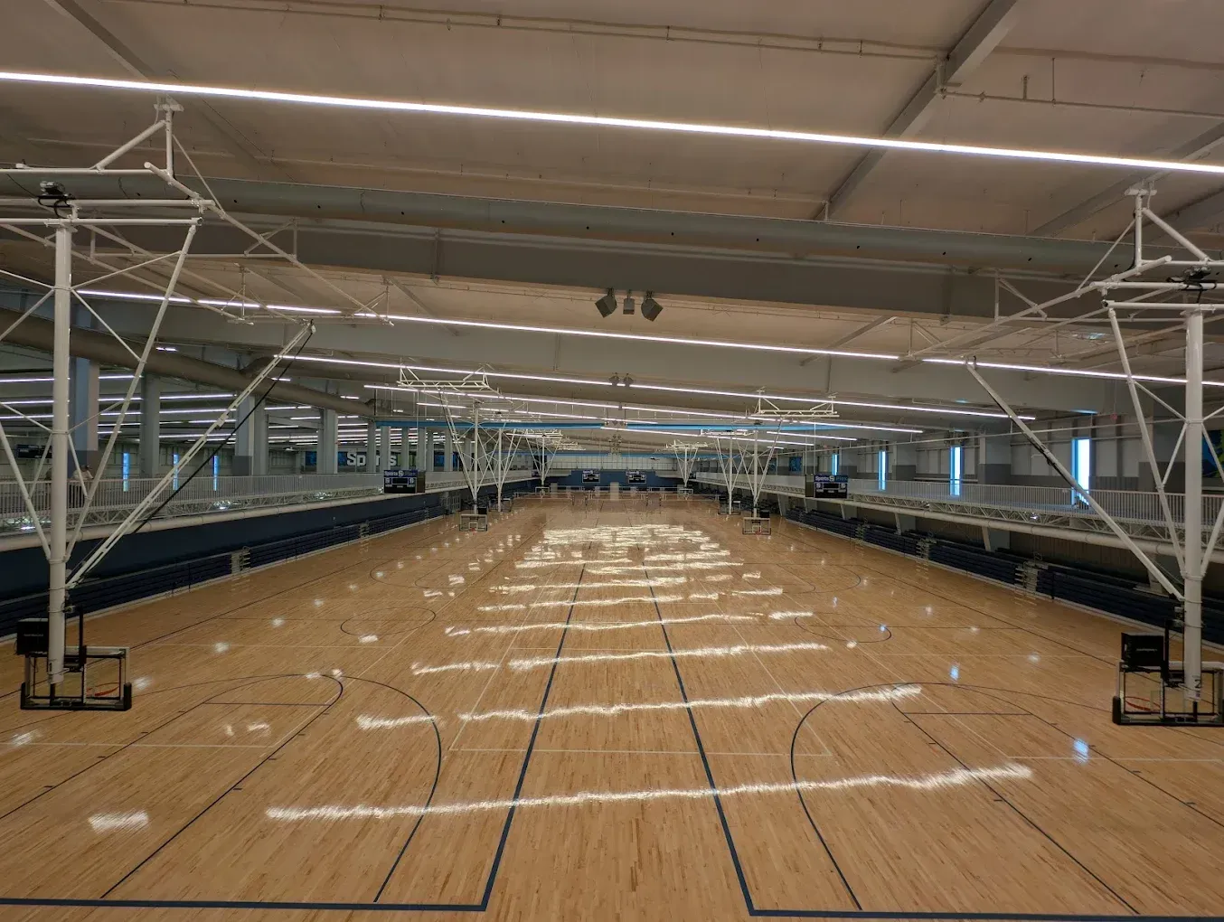 Empty indoor sports hall with a wooden floor and rows of blue seating under a high roof.