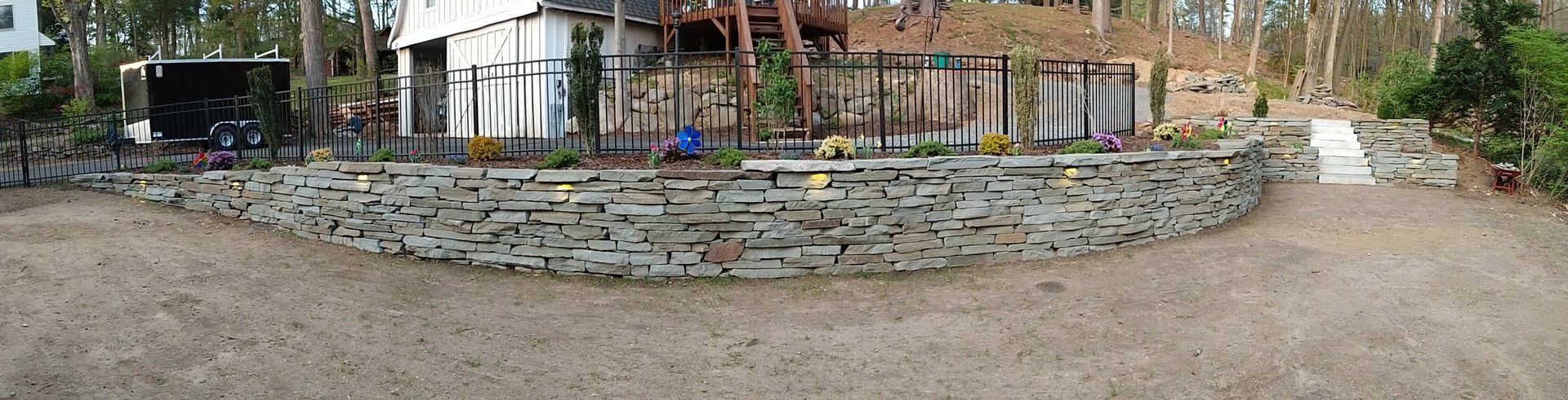Stone wall in front of a house, with a gravel driveway. Trees surround the property.