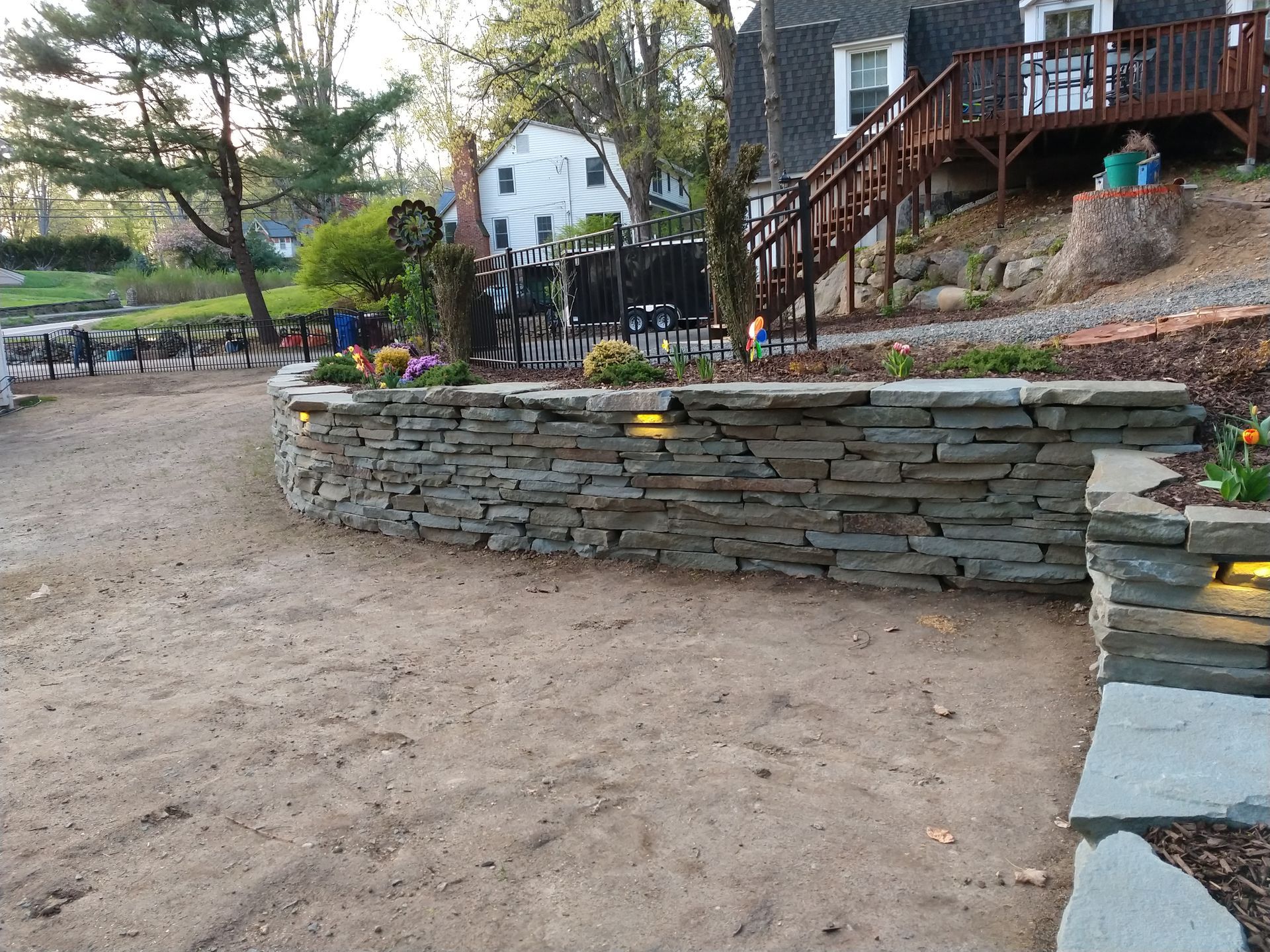 Stone retaining wall with built-in lights and planted flowers, gravel path, and house with wooden deck in background.