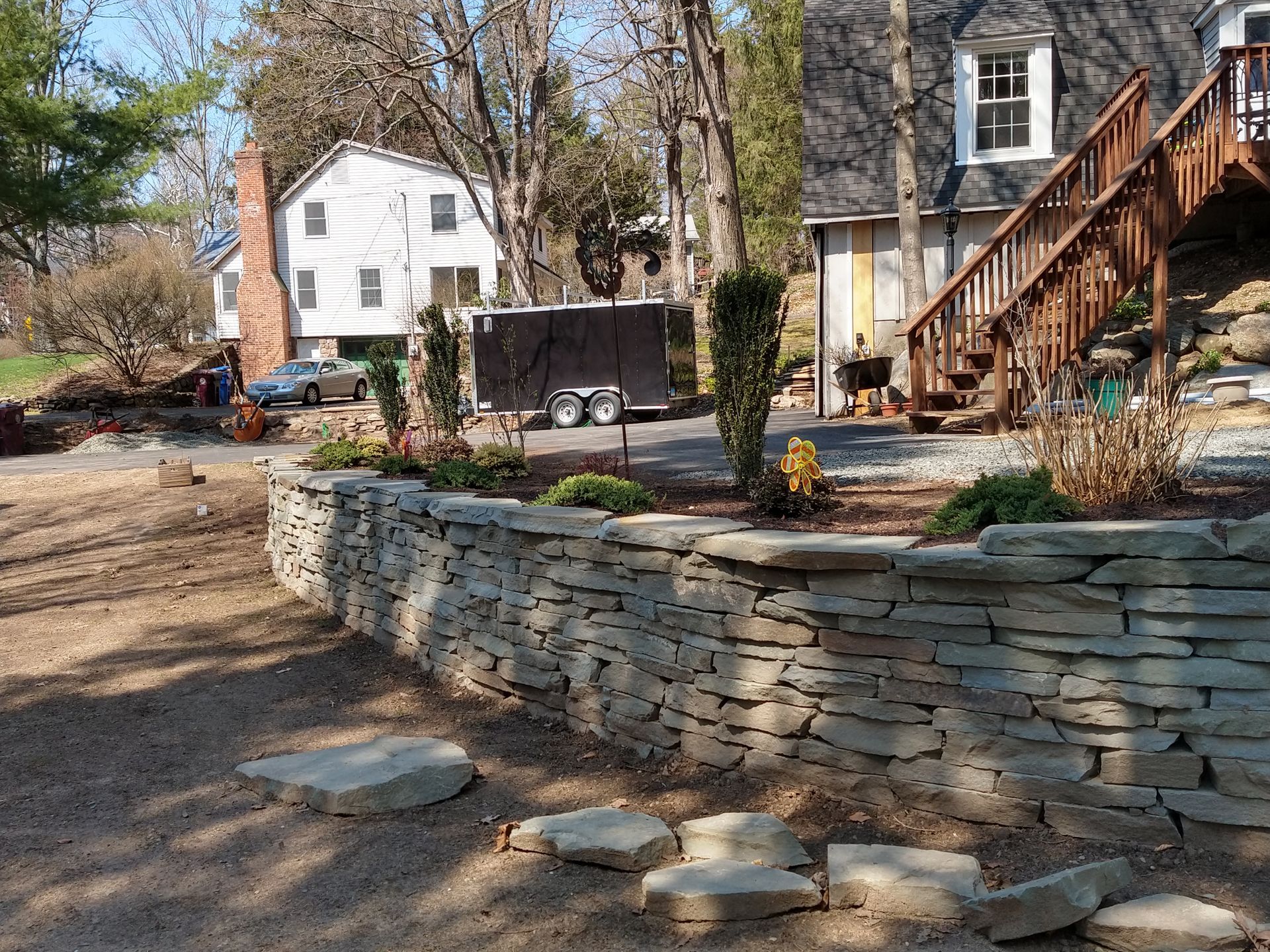 Stone retaining wall in a yard, with stepping stones. Houses and a trailer are in the background.