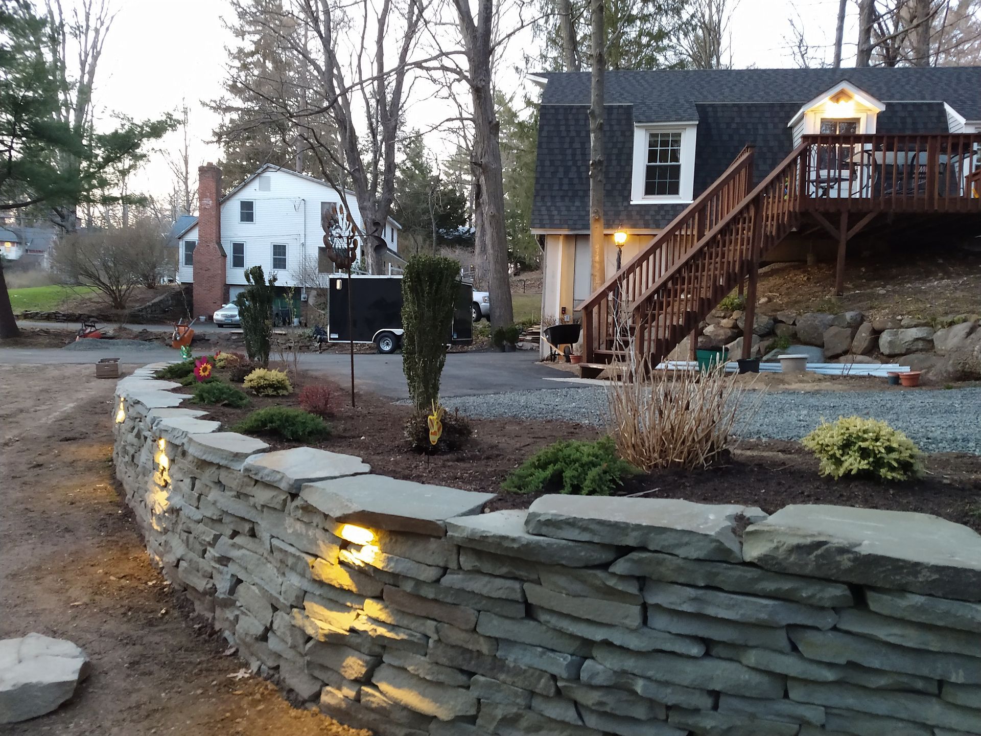 Stone retaining wall with built-in lights, landscaping, and a house with a deck in the background.