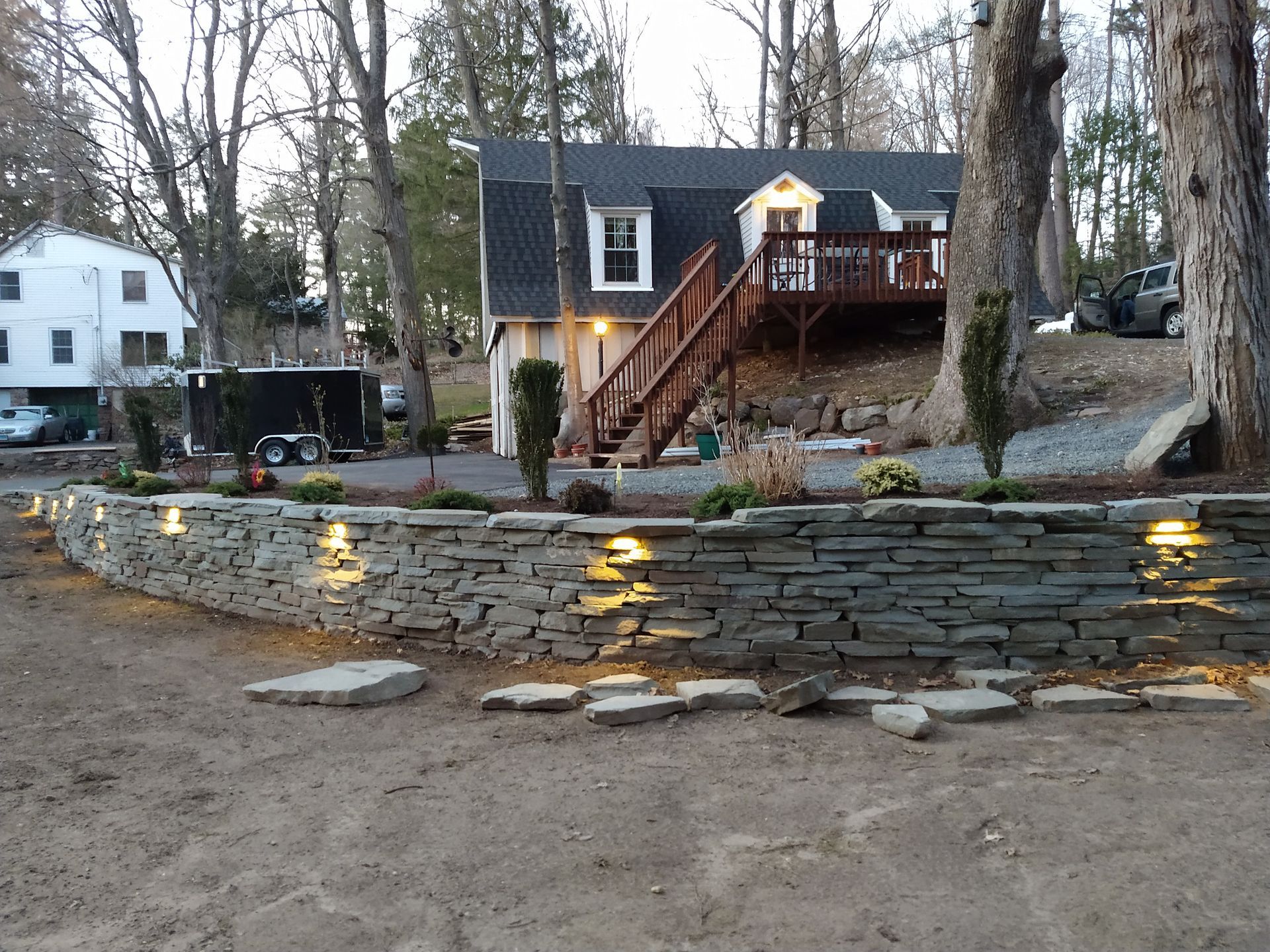 Stone retaining wall with built-in lights in front of a house with a deck and a dark roof.